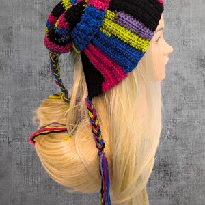Può includere: Un cappello colorato all'uncinetto fatto a mano con un motivo a righe in nero, rosa, blu, giallo e viola. Il cappello ha lunghe nappe intrecciate in colori coordinati. Il cappello è esposto su una testa di manichino.