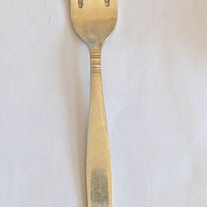 Puede incluir: Un tenedor plateado con cuatro púas, un mango largo y plano y un diseño ornamental decorativo en la base. El tenedor es de metal brillante y está sobre un fondo blanco. Este utensilio es adecuado para comer.