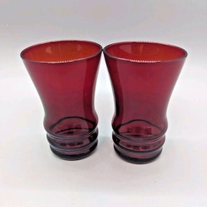 May include: Two ruby red drinking glasses. The glasses have a flared top and a curved, ridged base. The interior of the glasses is a lighter shade of red. The glasses are set against a white background.