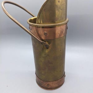 May include: A vintage brass coal scuttle with a copper band and handle. The scuttle has a curved top and a cylindrical body. The handle is arched, and the copper band is riveted to the brass. The scuttle is designed to hold coal.