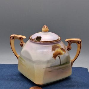 May include: A square porcelain sugar bowl with two handles and a lid featuring a hand-painted landscape with a tree. The bowl has a soft iridescent finish with yellow and tan accents. A copper-colored U.S. penny is placed in front for scale.