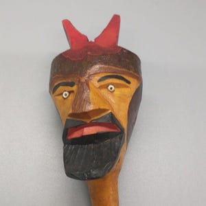 May include: A wooden carved figure of a person's head with a red hat and a black beard. The figure has a yellow face with painted white eyes.
