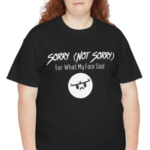 May include: Black t-shirt with the text "SORRY (NOT SORRY) For What My Face Said" in white. The shirt features a white circle with a sad face graphic. The model is wearing blue jeans.