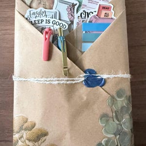 May include: A brown paper package tied with twine, sealed with blue wax, and decorated with floral stickers and illustrations. The package contains stationery, pens, and a book review card. Text includes "BOOK REVIEW" and "EASILY SLEEP IS GOOD."