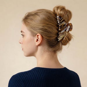 Puede incluir: El cabello de una mujer está recogido en un moño, sujeto con una pinza grande con estampado de carey. La pinza presenta tonos marrones, beige y crema. Lleva un suéter azul marino acanalado sobre un fondo neutro.