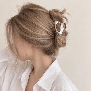 Puede incluir: Una pinza de pelo de metal plateado brillante con una forma irregular, orgánica y fluida. La pinza está sujeta en el cabello castaño claro peinado en un moño suelto, con una camisa blanca de cuello visible en la parte inferior.