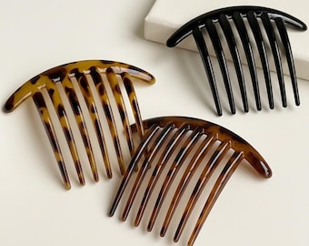 Puede incluir: Tres peines para el cabello con partes superiores curvas y múltiples dientes. Un peine es de color negro sólido, mientras que los otros dos presentan un patrón de carey marrón y negro. Los peines están dispuestos en grupo, mostrando su acabado.