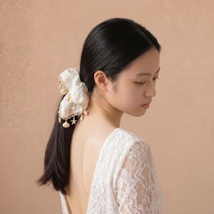 May include: A woman with long black hair, wearing a white lace dress, and a white hair accessory adorned with gold-colored stars and shells. The background is a solid, light brown color.