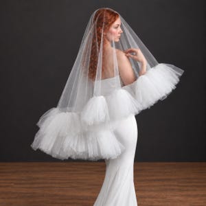 May include: A white bridal veil featuring a sheer, lightweight tulle fabric with a voluminous, multi-layered ruffled trim along the bottom edge. The veil is draped over the head and shoulders, extending outward to showcase the gathered tulle texture.