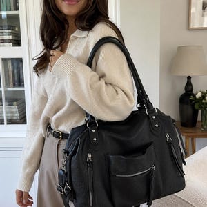 Op de afbeelding: Een zwarte leren handtas met meerdere vakken met ritssluiting en een schouderriem. De tas heeft een gestructureerd oppervlak en zilverkleurige hardware. Het heeft een voorvak en decoratieve kwastjes op de ritsen. De tas wordt door een persoon gedragen.