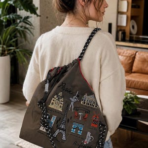Könnte beinhalten: Ein brauner Kordelzugbeutel mit einem schwarz-weißen Seilriemen. Die Tasche zeigt gestickte Bilder von Pariser Wahrzeichen, darunter der Eiffelturm und die Texte "Paris" und "Cafe/Bistro". Die Tasche hat ein rotes Innenfutter.