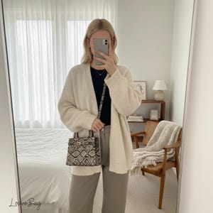Puede incluir: Una mujer con un cárdigan crema sostiene un bolso pequeño. El bolso tiene un estampado de piel de serpiente y una correa negra. Lleva una camiseta negra y pantalones grises. El espejo refleja un dormitorio con una cama.