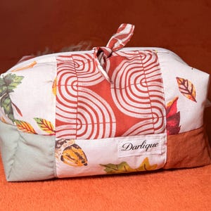 May include: A rectangular fabric pouch featuring a patchwork design with autumn-themed prints of leaves and acorns in shades of orange, brown, and green. The center panel displays a geometric red and white pattern. A label reads Darlique.