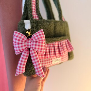 May include: A dark olive green textured fabric handbag featuring a pink and white gingham patterned ruffle trim and matching shoulder straps. A detachable pink and white gingham fabric bow is attached to the side with a gold metal clasp.