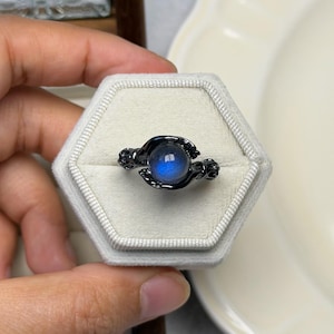 May include: A black ring featuring a blue gemstone, presented in a white, hexagonal ring box. The ring's design is decorative, and the gemstone is spherical. The ring is shown in a hand.
