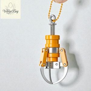 May include: A miniature claw machine pendant with a silver claw and orange and silver body. The claw is suspended from a chain. The pendant is held by a hand against a white background. The logo "Velora Bag" is in the upper left corner.