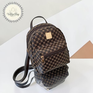 Può includere: Un mini zaino marrone e dorato fantasia con manico superiore, tasca frontale con cerniera e spallacci regolabili. La borsa ha un accento quadrato color oro sul davanti. La borsa è appoggiata su una superficie riflettente.