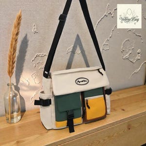 Puede incluir: Un bolso mensajero multicolor con una correa para el hombro negra. El bolso presenta una solapa de color crema con la palabra "Aputte" y dos bolsillos delanteros verdes y amarillos. También se ve un bolsillo lateral y una hebilla negra.