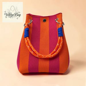 Può includere: Borsa tote a righe arancioni e magenta. Ha manici in corda con dettagli blu e finiture in metallo argentato. La borsa ha una chiusura con bottone e un logo nell'angolo in alto a sinistra. Un accessorio alla moda.