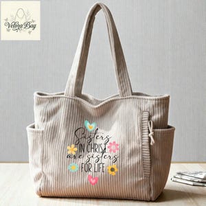 May include: Beige corduroy tote bag with two exterior pockets and long handles. The bag features the text "Sisters in Christ are sisters for life" surrounded by colorful floral and heart graphics. The Velora Bag logo is in the upper left corner.