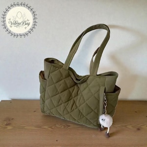 Peut inclure: Sac fourre-tout matelassé vert olive avec deux poches extérieures et une poignée assortie. Le sac présente un motif en losange et un petit porte-clés duveteux. Le sac est posé sur une surface en bois. Le nom de la marque "Velora Bag" est dans un logo circulaire.