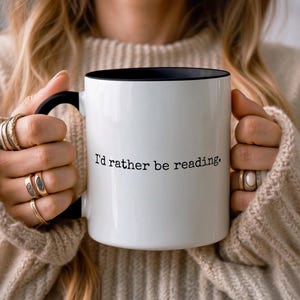 Peut inclure: Mug en céramique blanche avec intérieur et anse noirs. Le mug porte l'inscription "I'd rather be reading" en noir. La tasse est tenue par une personne portant un pull beige et plusieurs bagues.