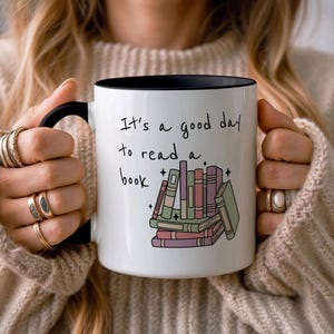 Peut inclure: Mug en céramique blanche avec intérieur et anse noirs. Le mug porte l'inscription "It's a good day to read a book" avec une pile de livres colorés. Le mug est tenu par une personne portant un pull beige.