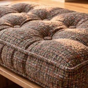 May include: A tufted cushion with a textured, multi-colored fabric in shades of brown, green, and orange. The cushion is rectangular and features button detailing. It rests on a wooden surface, suggesting a piece of furniture.