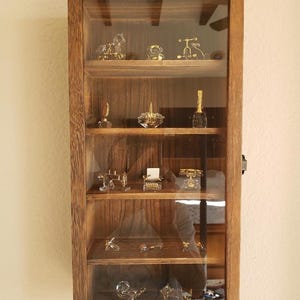 May include: A rectangular wooden display case with a glass front door and five interior shelves. The cabinet features a natural wood grain finish and contains various small gold-toned and clear glass miniature figurines arranged on the shelves.