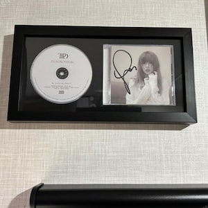 May include: A black rectangular frame containing a white compact disc labeled TTPD and a square album cover featuring a black ink autograph over a portrait of a person with long hair wearing a white shirt, mounted on a black background.