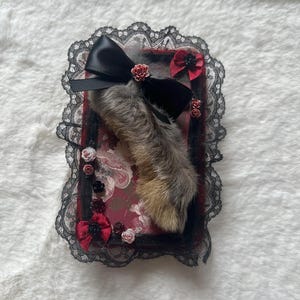 May include: A rectangular decorative plaque featuring a central grey and brown animal paw, adorned with a black satin bow, black lace trim, and small red, black, and white fabric roses. The background is a dark red floral patterned surface.