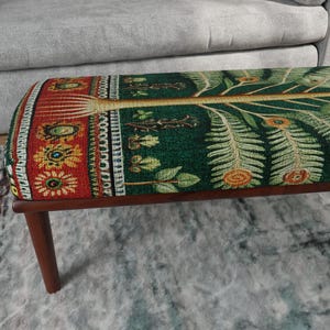 Puede incluir: Un banco rectangular con patas de madera marrón oscuro y un asiento acolchado tapizado en tela verde con un motivo de árbol detallado, hojas tipo helecho, acentos circulares naranjas y un borde rojo con patrones florales.