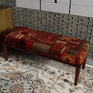 Könnte beinhalten: Eine rechteckige Bank mit einer gepolsterten Sitzfläche, die mit einem mehrfarbigen Stoff im Patchwork-Design aus roten, orangefarbenen und braunen geometrischen Mustern bezogen ist. Die Bank steht auf vier dunkelbraunen Holzbeinen.