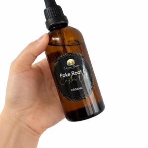 May include: A brown glass bottle of Organic Root & Castor Oil with a black dropper top. The bottle has a black label with white text that reads "Poke Root & Castor Oil" and "Organic". The bottle is held in a hand.