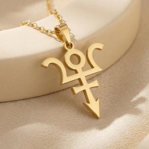 May include: Gold-colored pendant necklace featuring a stylized symbol. The pendant has a unique design with curved and pointed elements. The necklace chain is delicate and complements the pendant's design. The jewelry is displayed on a neutral background.