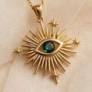 May include: Gold-colored necklace featuring a pendant with an eye design. The eye has a green gemstone center, surrounded by a sunburst design with star accents. The necklace is displayed on a cream-colored fabric.