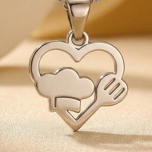 May include: Silver-toned pendant necklace featuring a heart-shaped design with a chef's hat and spoon. The pendant hangs from a delicate chain. The necklace is displayed on a light-colored surface.