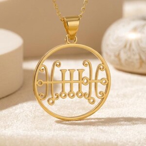 May include: A gold pendant necklace with a circular pendant and an intricate, stylized design. The pendant is suspended from a fine chain. The necklace is presented on a cream-coloured surface.