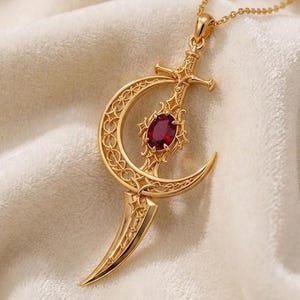 May include: A gold-toned necklace featuring a pendant shaped like a curved sword intersecting a crescent moon. The pendant is decorated with intricate filigree patterns and a single oval-cut deep red gemstone set in the center of the sword hilt.