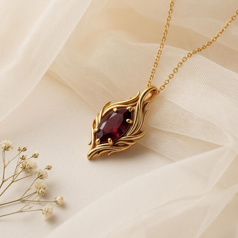 May include: A gold-colored pendant necklace featuring a large, oval, deep red gemstone. The pendant has an ornate, stylized leaf design surrounding the stone. The necklace is displayed on a cream-colored fabric.