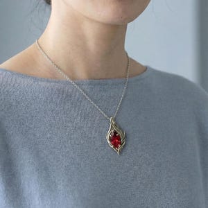 May include: A silver necklace with a pendant featuring a vibrant red gemstone. The pendant has a stylized, flame-like design. The necklace is worn against a light blue top.