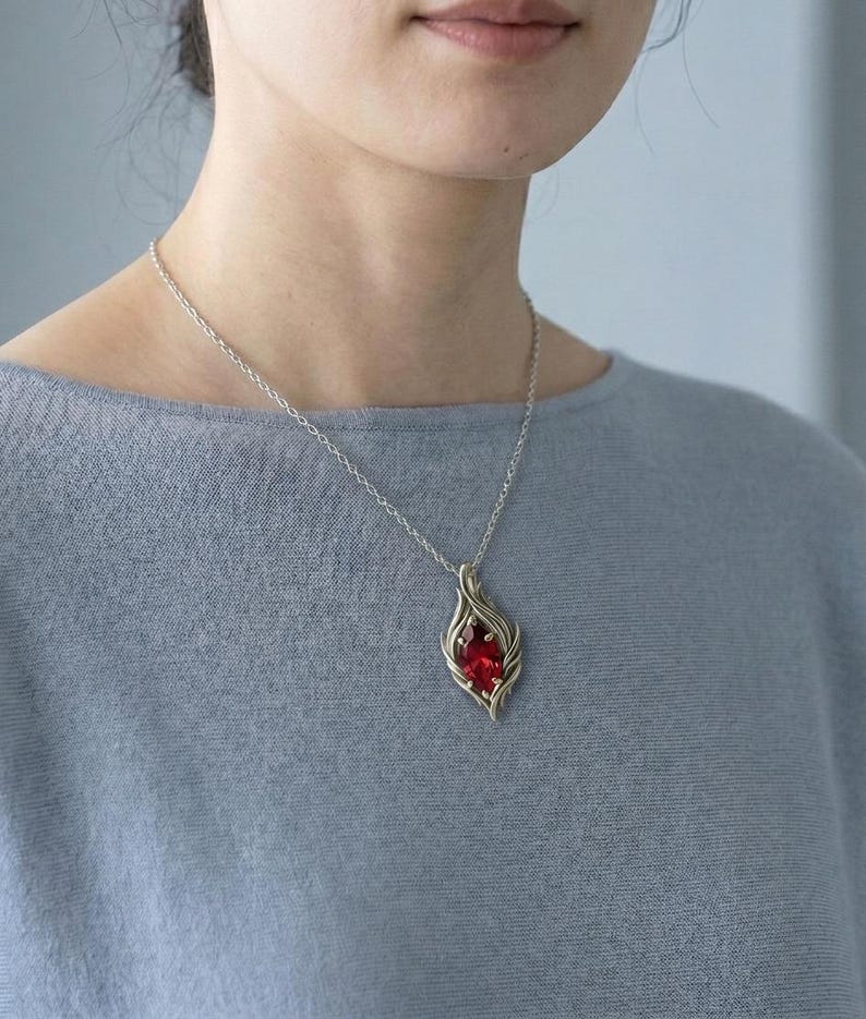 May include: A silver necklace with a pendant featuring a vibrant red gemstone. The pendant has a stylized, flame-like design. The necklace is worn against a light blue top.