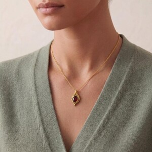 May include: A gold-colored pendant necklace featuring a teardrop-shaped, dark red gemstone. The pendant has an ornate, openwork design. The necklace is worn against a light green sweater.