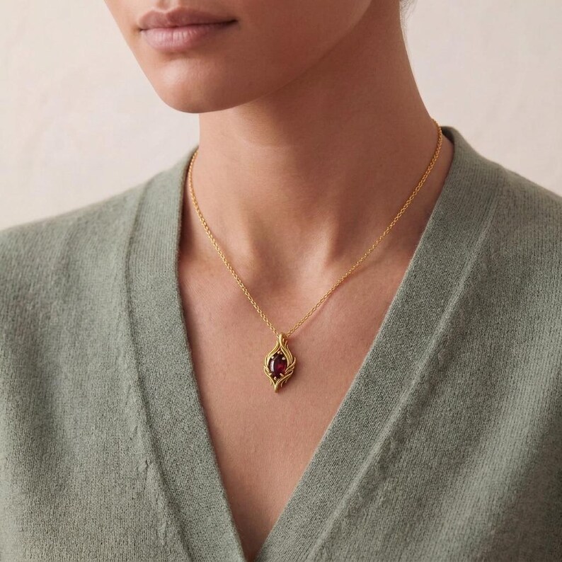 May include: A gold-colored pendant necklace featuring a teardrop-shaped, dark red gemstone. The pendant has an ornate, openwork design. The necklace is worn against a light green sweater.