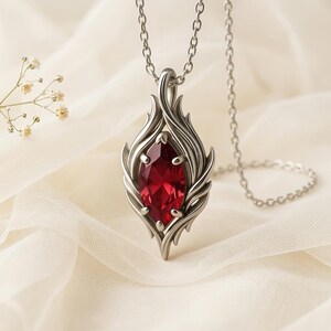 May include: A silver pendant necklace featuring a large, faceted, red gemstone. The pendant has a stylized flame design around the gem. The necklace chain is also silver and hangs on a white fabric background.
