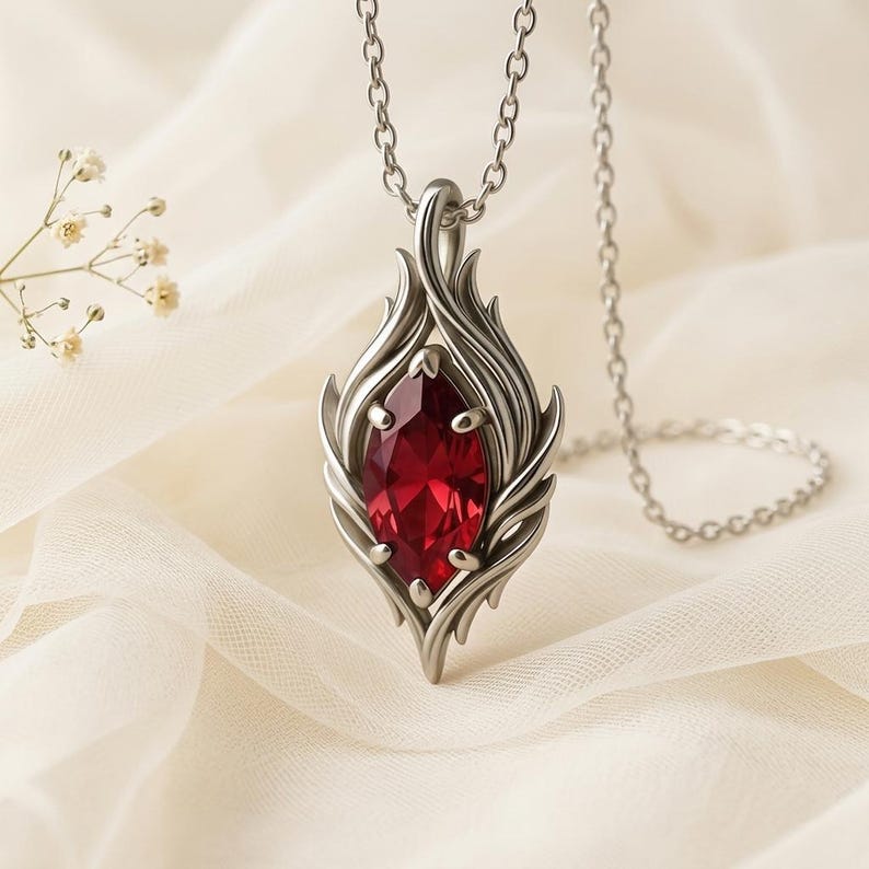 May include: A silver pendant necklace featuring a large, faceted, red gemstone. The pendant has a stylized flame design around the gem. The necklace chain is also silver and hangs on a white fabric background.