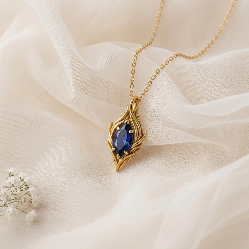 May include: A gold-colored pendant necklace with a large, oval, deep blue gemstone. The pendant is encased in a stylized flame design. The necklace chain is delicate, and the background is a soft, white fabric.