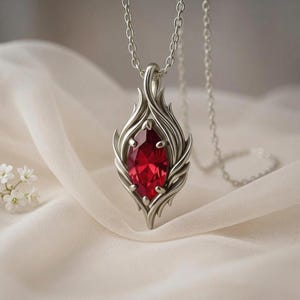 May include: A silver pendant necklace featuring a large, oval-cut, red gemstone. The gemstone is set within a stylized, flame-like silver frame, suspended from a delicate chain. The design is elegant and eye-catching.
