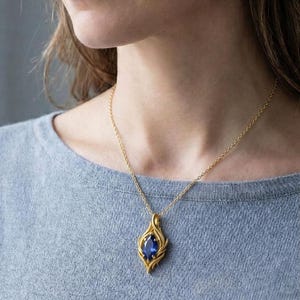 May include: A gold pendant necklace featuring a large, oval, deep blue gemstone. The pendant has a stylized, flame-like design. The necklace is worn on a person wearing a gray shirt.