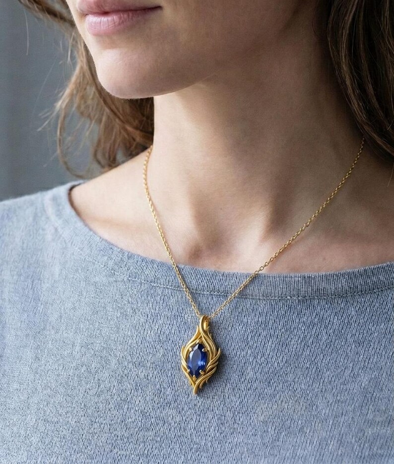 May include: A gold pendant necklace featuring a large, oval, deep blue gemstone. The pendant has a stylized, flame-like design. The necklace is worn on a person wearing a gray shirt.